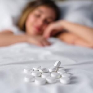 Close-up of white sleeping pills on a bed with a blurred woman sleeping in the background, representing sleep aid medication like Ambien or Zolpidem